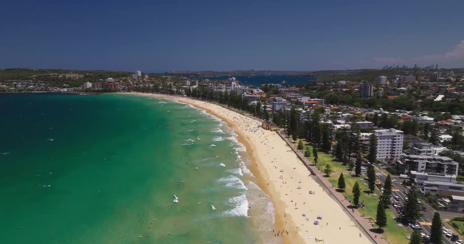 An aerial view of a coastal city featuring a long sandy beach with turquoise waters and numerous people enjoying the beach