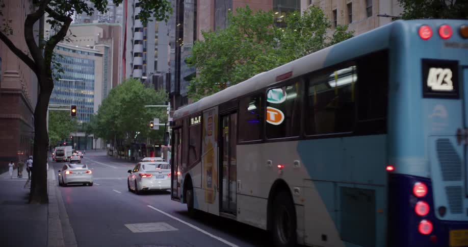 A white and blue bus drives down a city street lined with tall buildings alongside other vehicles under an overcast sky