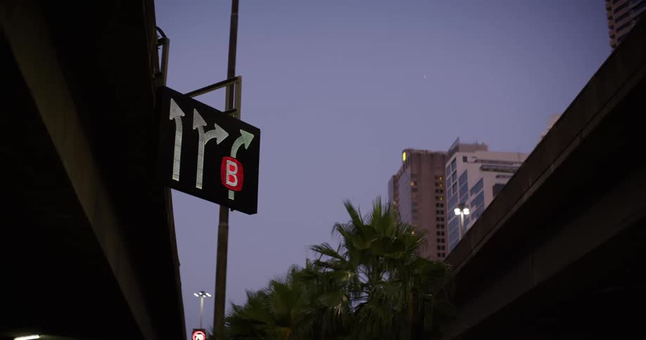A cityscape at dusk with a freeway overpass and a digital traffic sign displaying lane directions