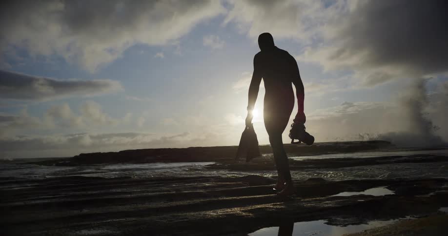 Filmmaker walking with a professional camera and swimming costume on a beach with waves crashing in the background