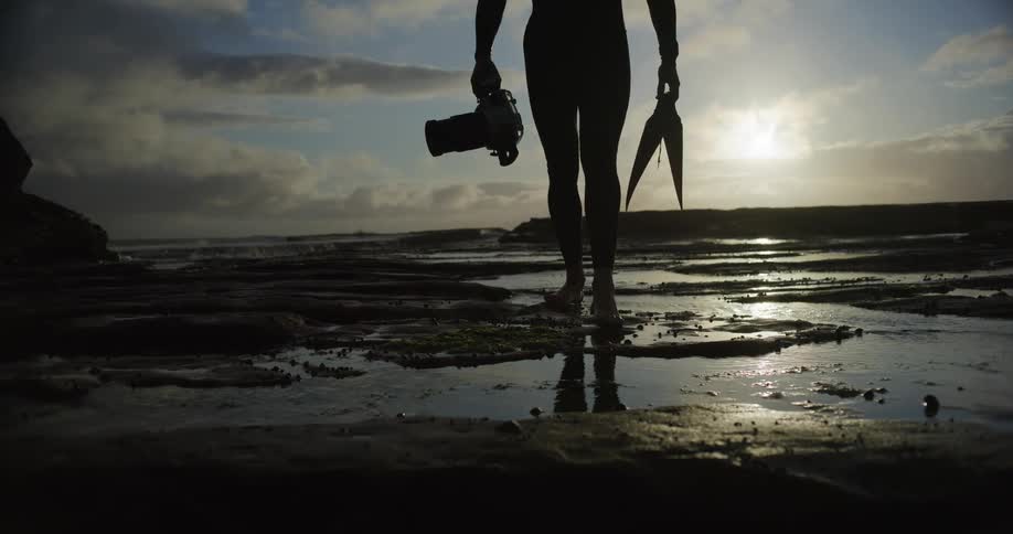 Silhouette of a person in a swimming suit walking along the beach carrying a professional camera