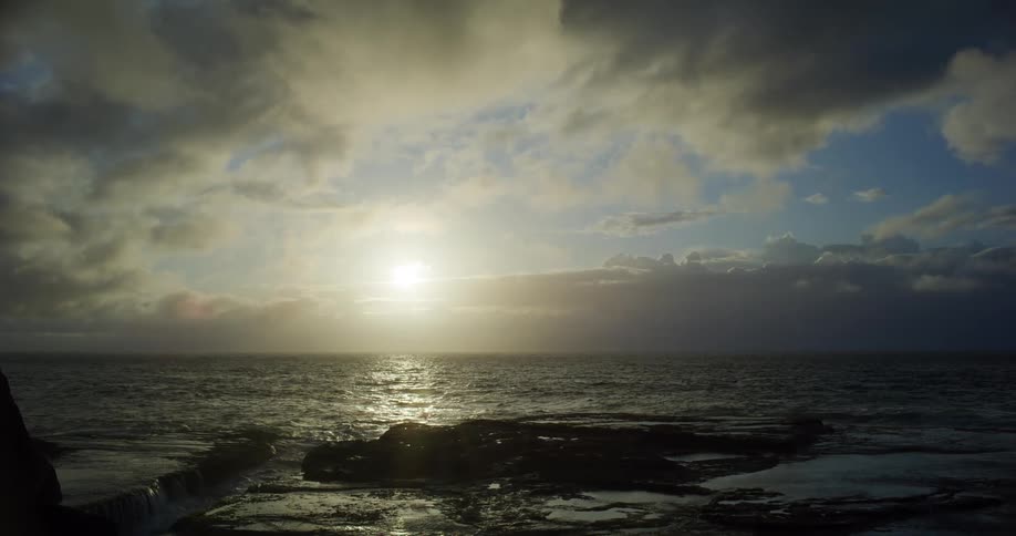 A rocky shoreline reflects the muted glow of a rising sun through a sky thick with clouds