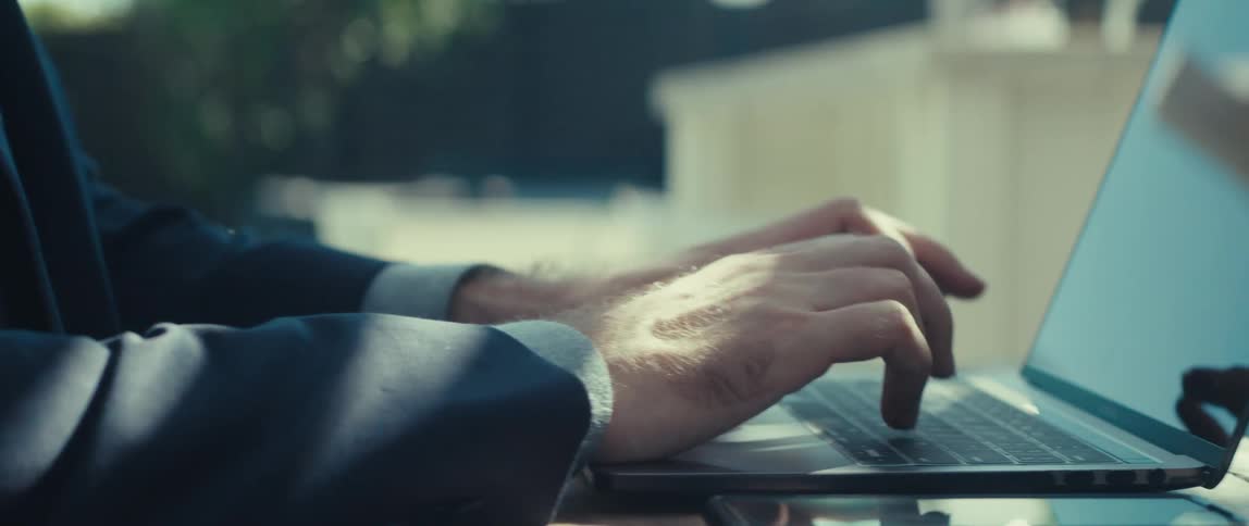 Man in blazer sitting typing on laptop and picking up his smartphone next to it