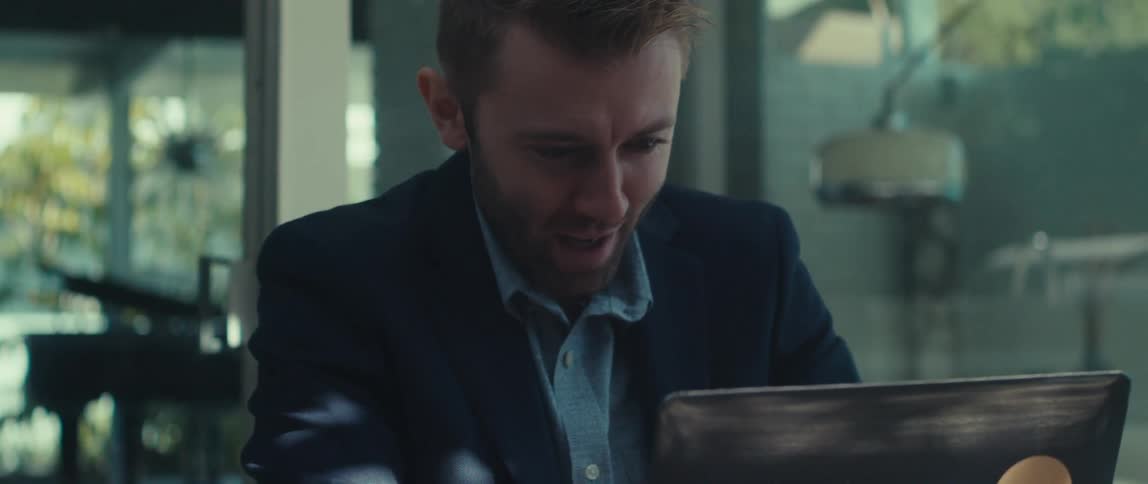 A man in a dark suit jacket and lightcolored shirt sits outdoors having a meeting on his laptop