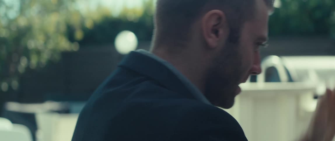 A man in a dark suit jacket and lightcolored shirt sits outdoors rising his arms in celebration