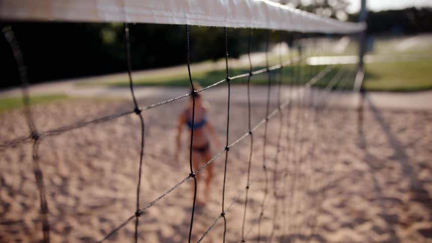 A volleyball net with its taut strings in focus while a beach volleyball player is out of focus in the background