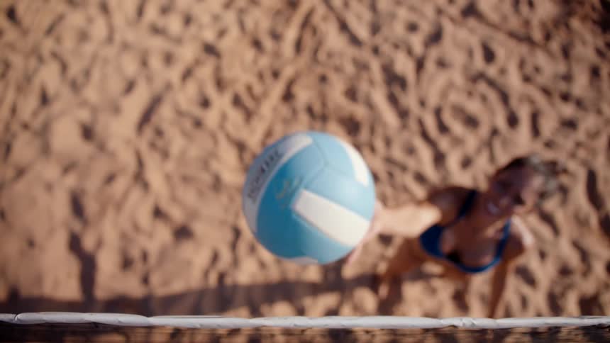 A smiling woman stands on a sand volleyball court and tosses a blue volleyball up into the air