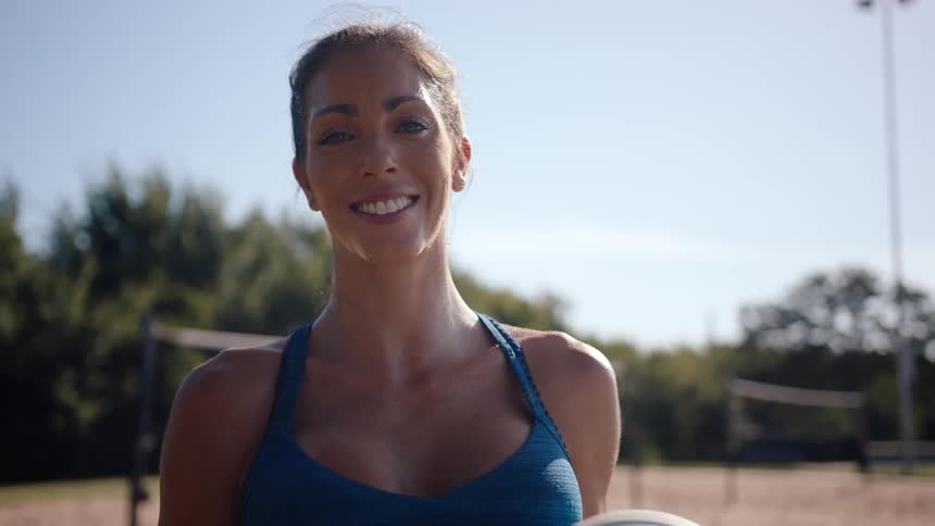 A woman holding a volleyball on a beach court smiling to the camera