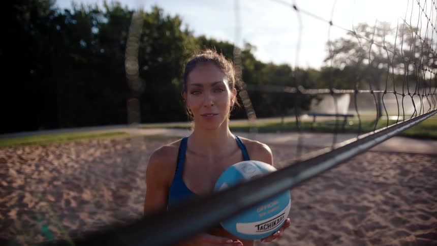 Woman holding a volleyball on a sand court with a sun on the horizon