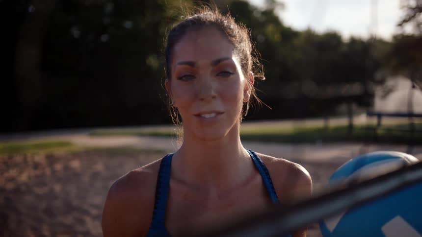 Portrait of a lighteyed woman smiling and holding a volleyball while standing on a sand court