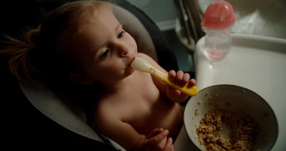 A toddler sits in her highchair eating a bowl of oatmeal with a plastic spoon