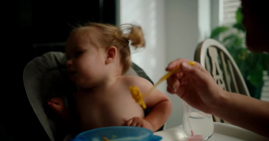 Toddler sitting in high chair refuses to eat food presented by her mother