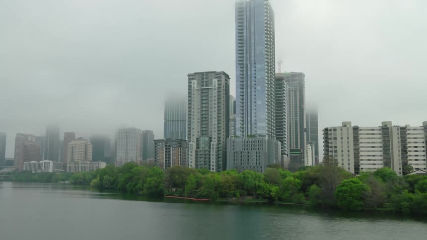 A misty cityscape with tall buildings is reflected in the calm waters of a lake