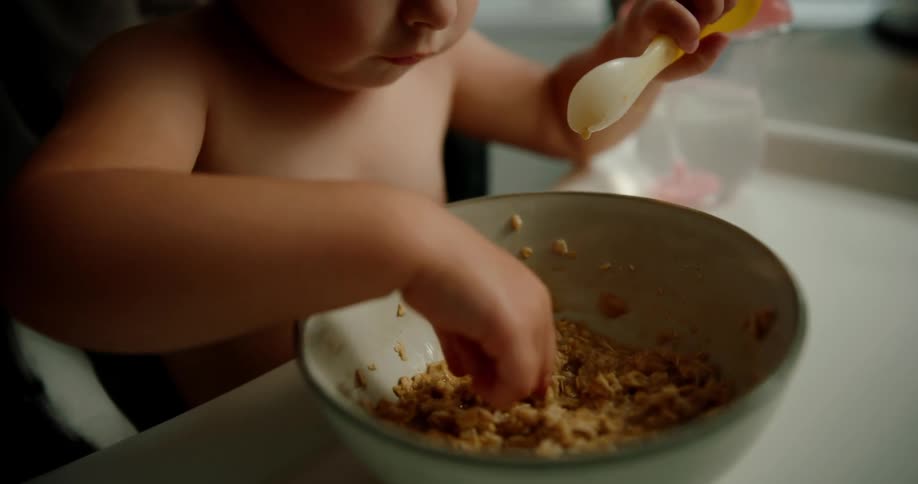 A young child using a yellow spoon to eat cereal from a bowl appearing focused on their meal in a softly lit indoor setting
