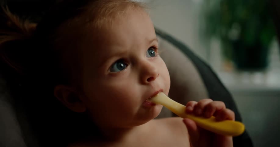 A young child is sitting in a high chair holding and nibbling on a spoon