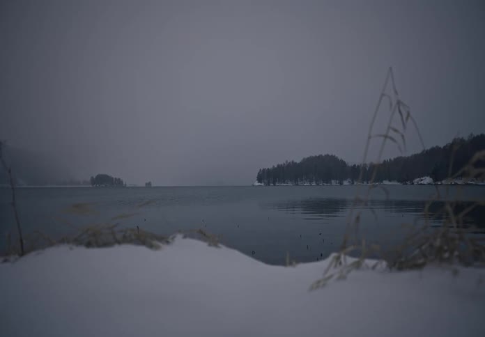 Snow covers the shores of a lake bordered by conifer covered hills on an overcast winter day