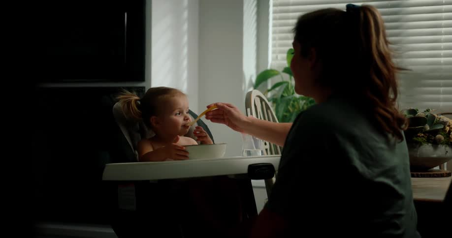 A woman is feeding a toddler sitting in a high chair with a spoon