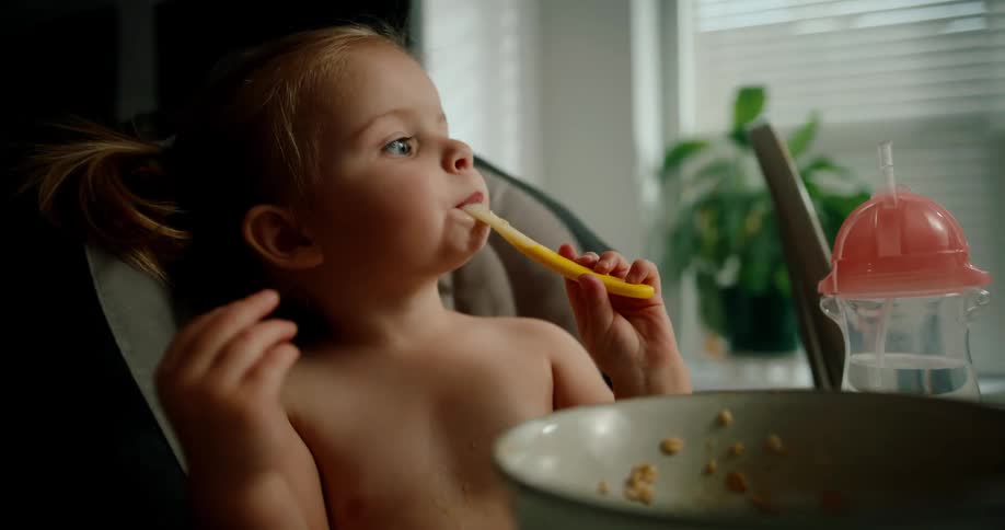 A young child is sitting in a high chair holding and nibbling on a spoon with a bowl and a sippy cup nearby