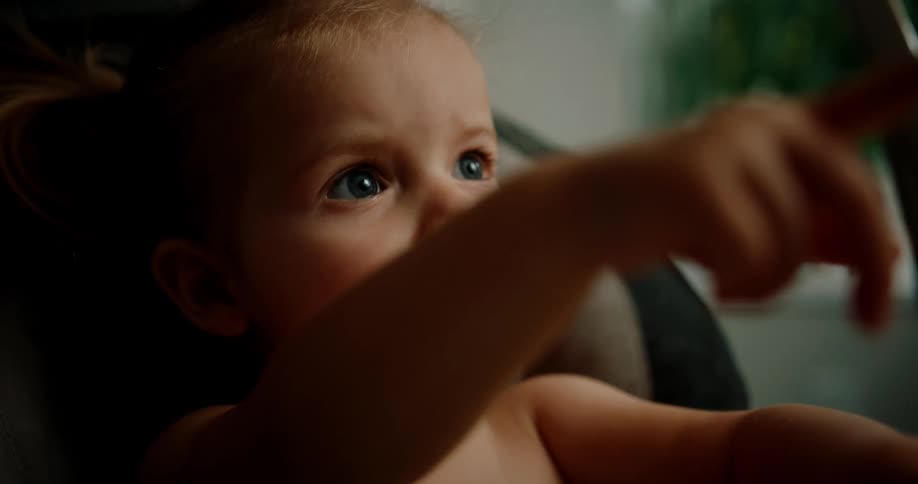 A young child sitting in a high chair is pointing with her finger