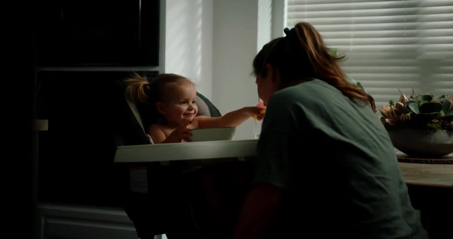 A young child sitting in a high chair smiles and offers a spoon to an adult leaning in with soft lighting illuminating the scene