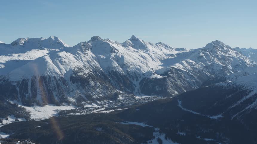 Aerial view of a snow capped mountain range with a charming village nestled in the valley below under a clear blue sky