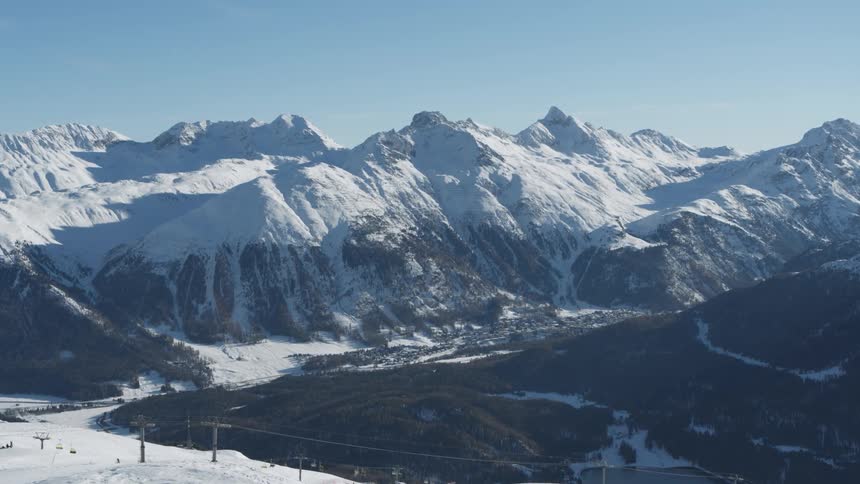 An aerial view of a snow covered ski resort with several ski lifts and slopes winding through the mountain landscape with a small town nestled in the valley in the distance near the mountain range