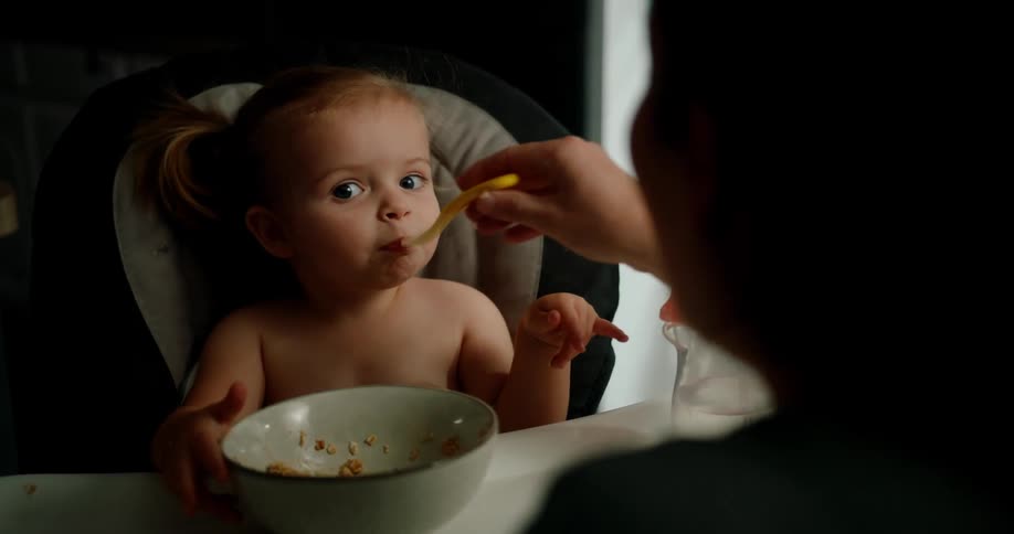 Child sitting in a high chair with a curious and playful look while being fed with a spoon