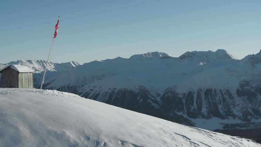 A winter scene at a ski resort with a small wooden hut a swiss flag waving proudly in the foreground and a view of snow capped mountains stretching towards the horizon