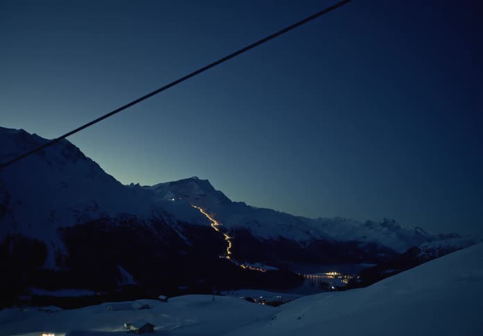 A nighttime view of a snowy mountain range lit by the winding trail of lights from a ski lift a small village below and a ski lift cable in the foreground