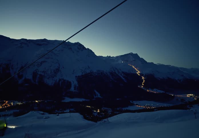 Moving view of a snowy mountain at night with cable cars and trails of city lights in the background