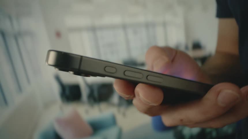 A slightly out of focus close up shows a persons hands holding and using a smartphone with a brightly lit office space blurred in the background