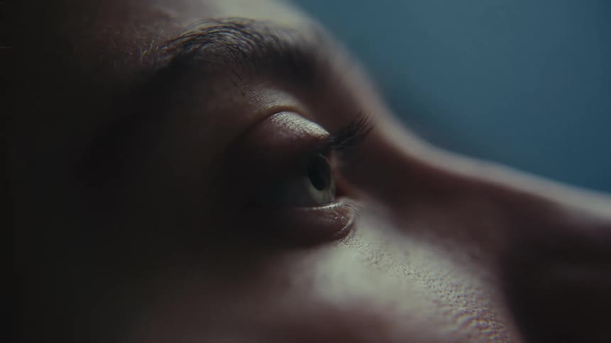 A close up shot gradually focuses on a persons eye revealing details of the eyelashes eyebrow and skin texture against a dark blue background