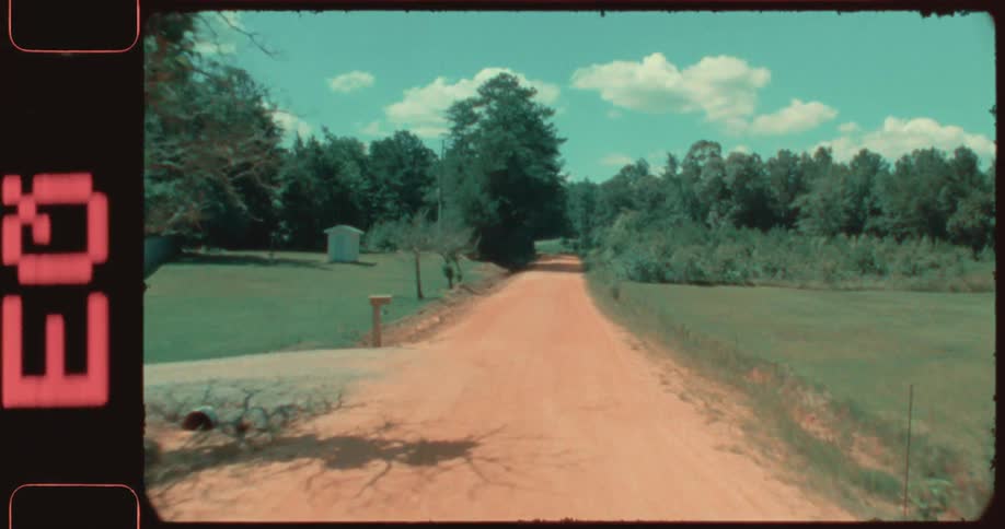 Red dirt road passes rural driveways and groves of trees on a cloudysummer day