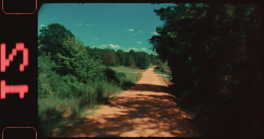 A vintage slightly shaky shot shows a dirt road stretching into the distance flanked by lush trees under a vibrant blue sky