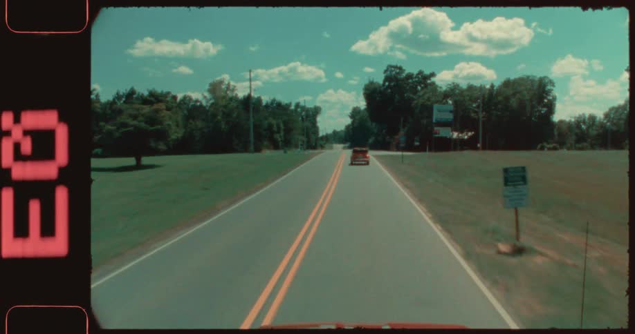 An orange convertible volkswagen beetle drives down a rural highway on a summer day