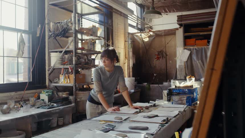 A woman with glasses is shown working at a cluttered table filled with papers in a sunlit industrial style studio space