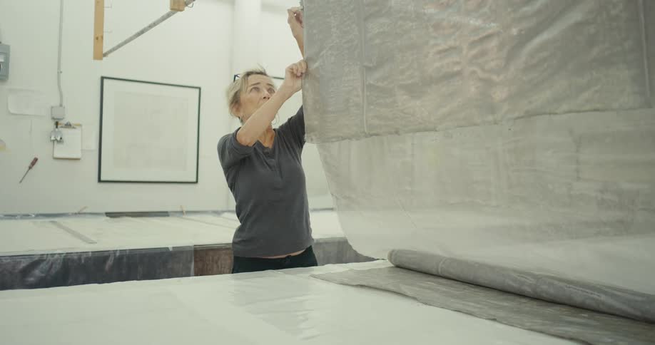 A woman is adjusting a large piece of fabric hanging from the ceiling over a large table in a workshop