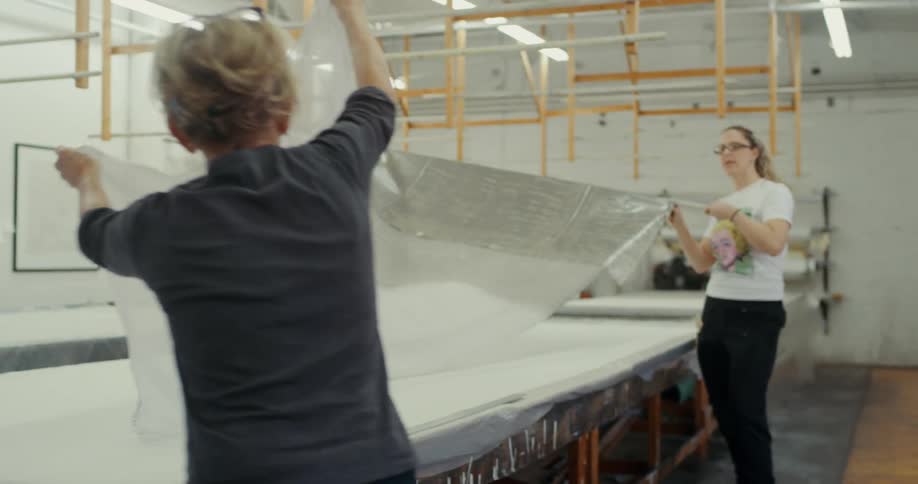 Three women in a workshop are shown working to unfurl a large sheet of semi transparent material over a long table