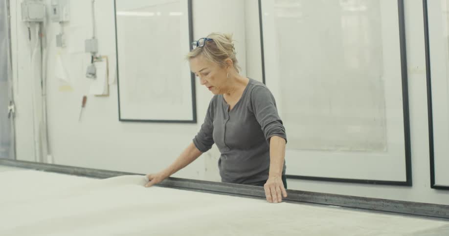 In a workshop a woman with glasses gently spreads a large white fabric across a table