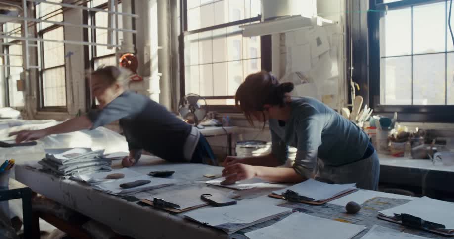 Two people working in a bright industrial style workspace with large windows papers and art supplies scattered across the tables