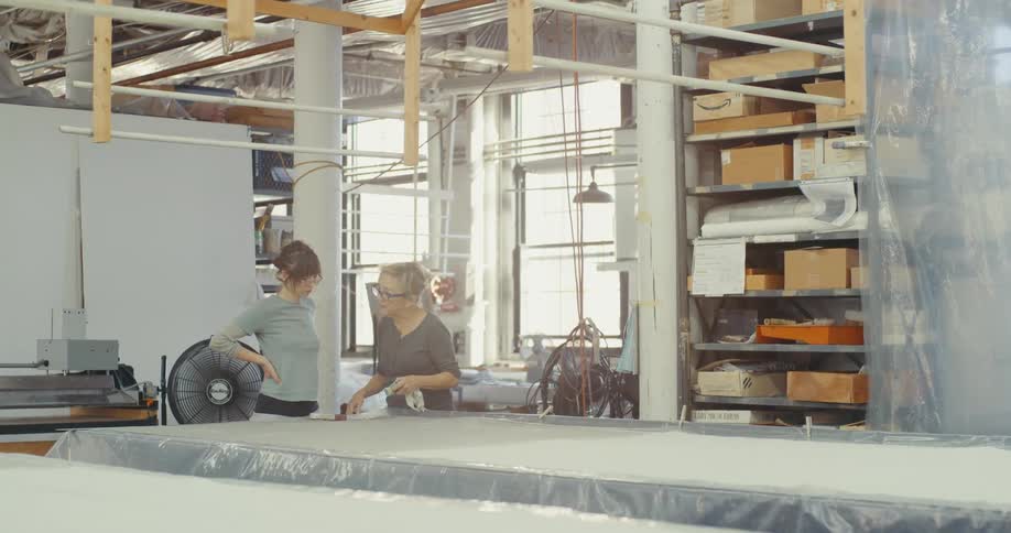 Two women in a well lit workshop one closely examining a large piece of material on a table while the other stands nearby with her hands on her hips observing the work