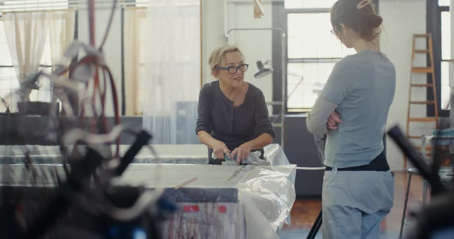 Two women in a workspace with one focused on cutting or measuring material on a table while the other stands nearby with arms crossed observing