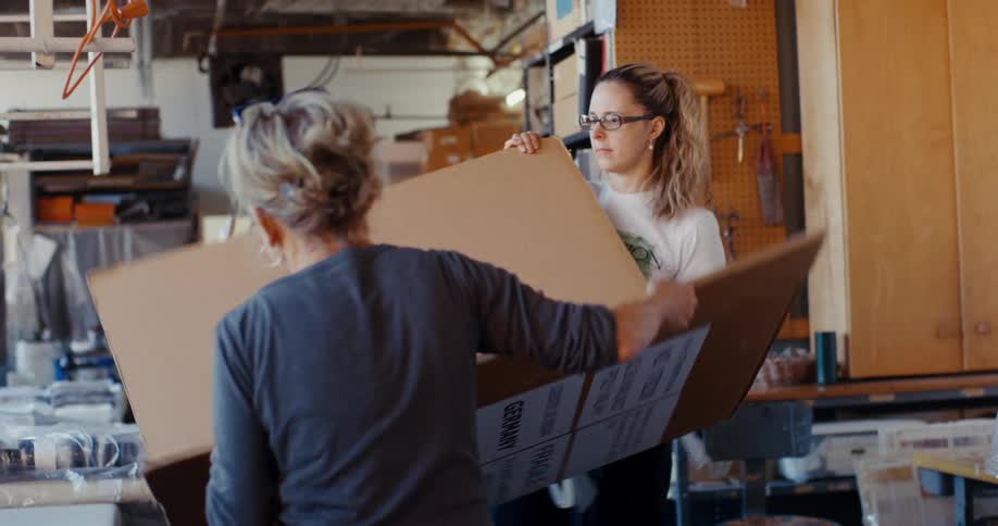 Two women in a well lit workshop place a large cardboard box on a table
