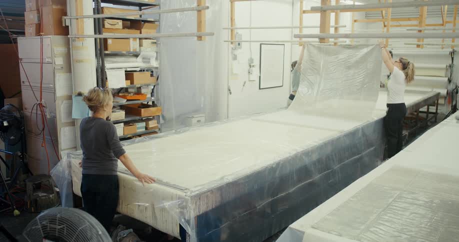 Three women in a workshop collaboratively handling a large sheet of transparent plastic material on a long table with shelves of boxes and supplies visible in the background