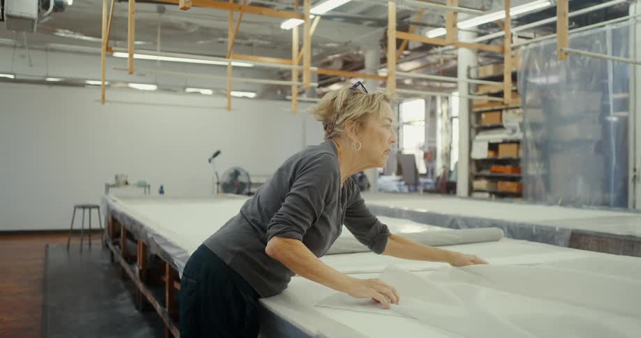 A woman in a gray shirt is spreading a large white sheet of fabric over a long table in a workshop space