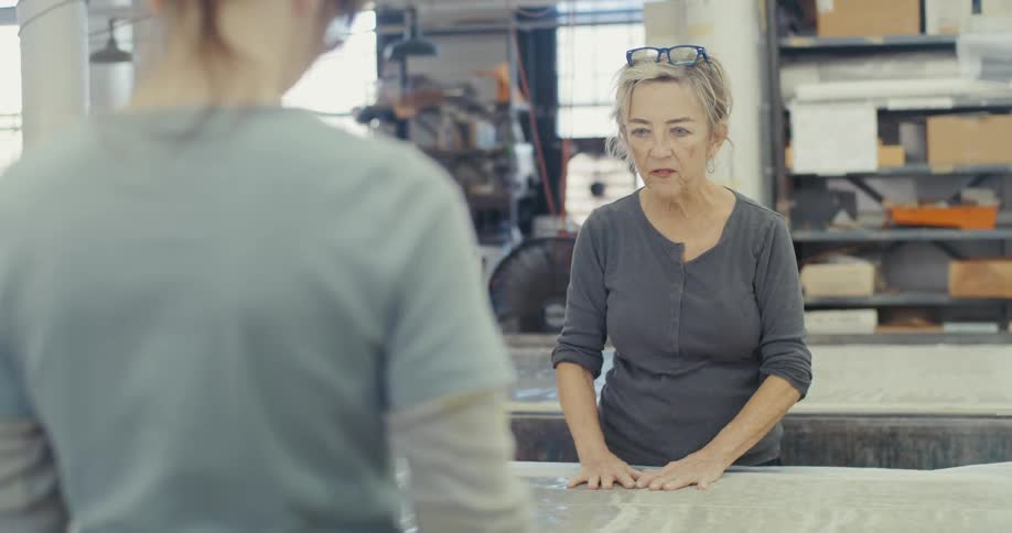 Two women are standing in a workshop engaged in a task involving a large table covered with a material