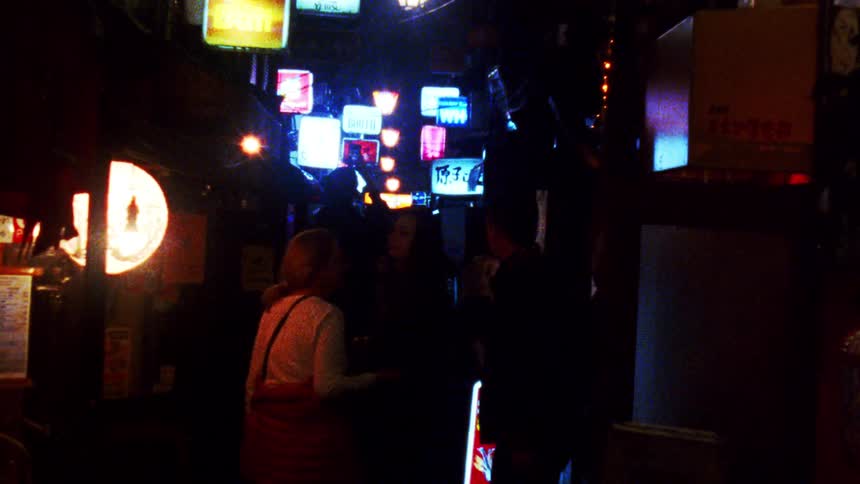 Neon signs hang in a dimly lit alleyway casting colorful shadows on people walking by