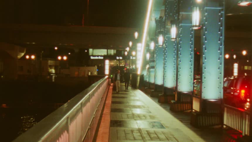 A nighttime scene of a welllit pedestrian walkway on a bridge with blue illuminated columns and a few people walking on it