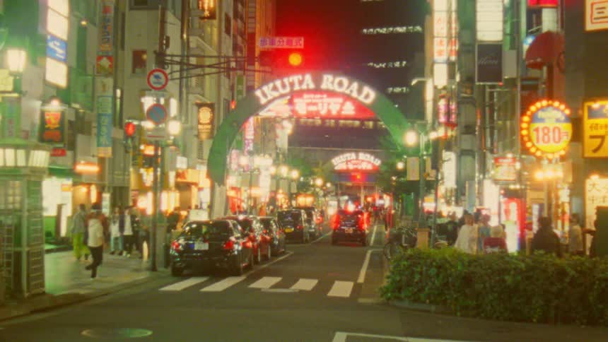 An asphalt street with some cars passing by and several pedestrians on the sidewalks and there is an arch with lights from illuminated signs above