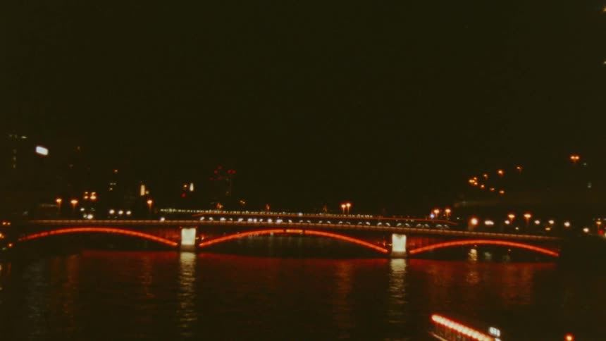 Bridge illuminated in vibrant shades of red reflects its lights on the dark waters of the river while a boat with soft lights glides silently